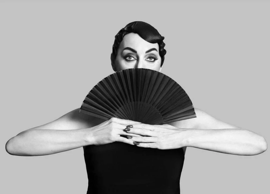 Black and white portrait of an adult female with a retro hairstyle, holding a black fan and posing against a grey background.