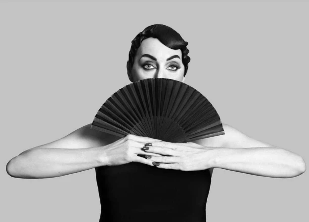 Black and white portrait of an adult female with a retro hairstyle, holding a black fan and posing against a grey background.