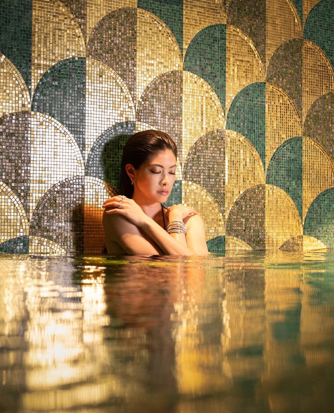 An Asian woman is shown from the chest up in water with a decorative tile wall behind her. The water reflects light and patterns.