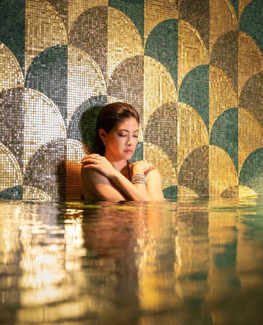 An Asian woman is shown from the chest up in water with a decorative tile wall behind her. The water reflects light and patterns.