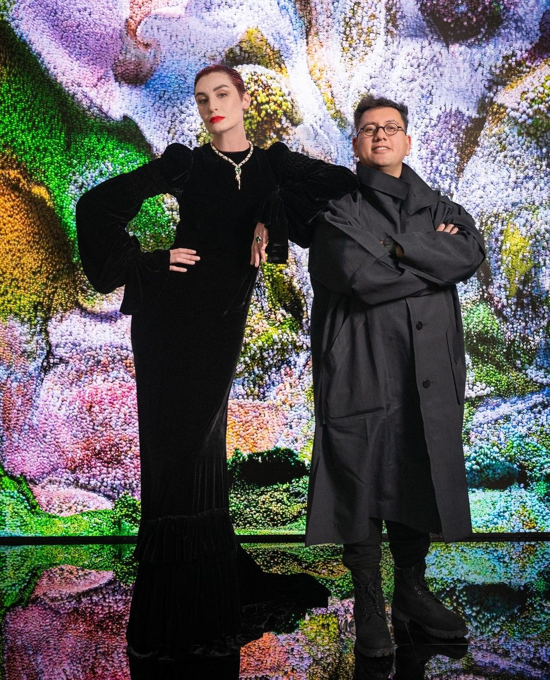 Two people stand in front of an abstract, colorful backdrop. A woman wears a long black velvet dress and jewelry. A man wears glasses and a black coat.
