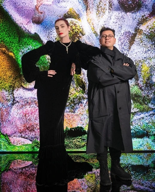 Two people stand in front of an abstract, colorful backdrop. A woman wears a long black velvet dress and jewelry. A man wears glasses and a black coat.