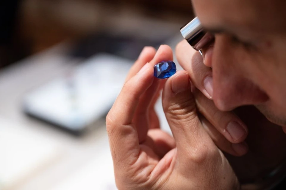 Close-up of a person inspecting a blue gemstone with a jeweler's loupe, highlighting the gem's facets and color under bright lighting.
