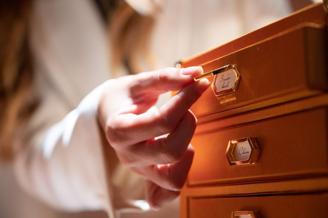 A woman's hand opens a wooden jewelry box drawer, revealing labels like 'snake head' and 'chain', bathed in soft, natural light.