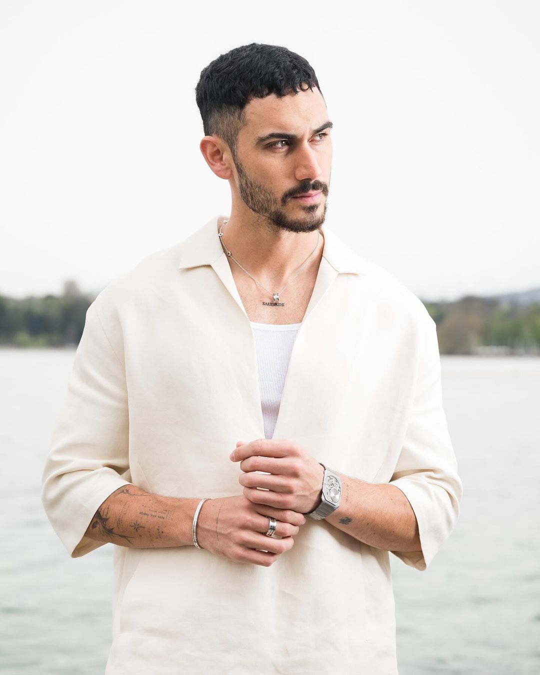 Portrait of adult male with short dark hair, wearing an open linen shirt, posing outdoors against a blurred background of lake and trees.