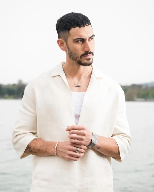 Portrait of adult male with short dark hair, wearing an open linen shirt, posing outdoors against a blurred background of lake and trees.