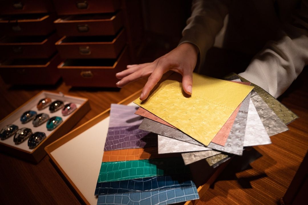 Person's hand displaying colorful leather swatches of varying textures on a wooden surface next to jewelry and drawers.
