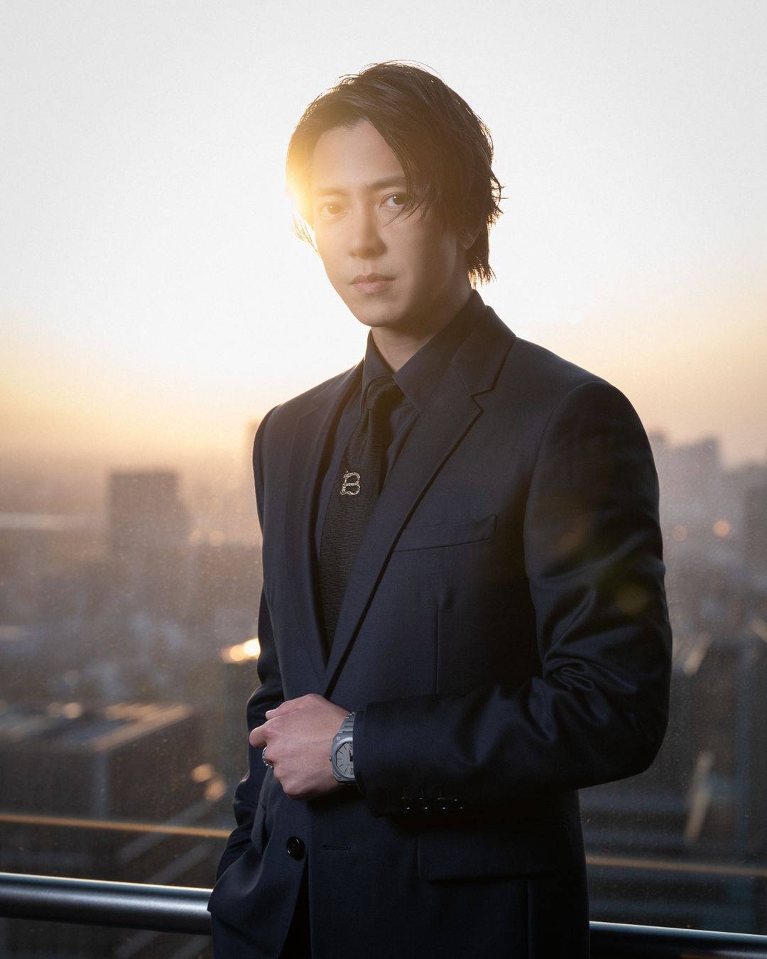 Portrait of an adult male in a dark suit with a serious expression and a wristwatch, posing outdoors with a cityscape in the background.