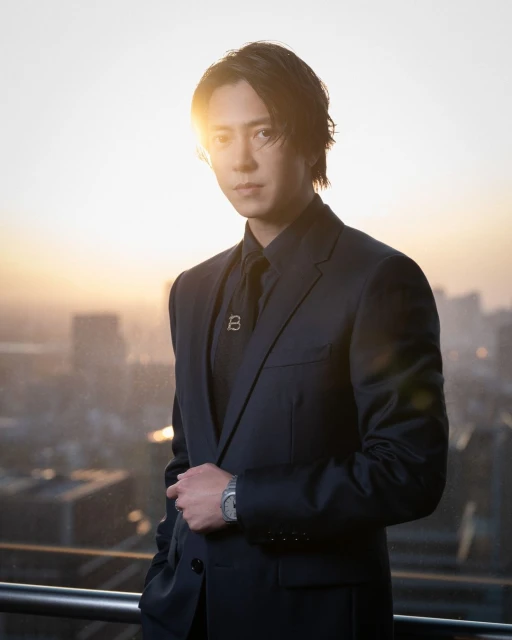 Portrait of an adult male in a dark suit with a serious expression and a wristwatch, posing outdoors with a cityscape in the background.