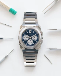 Overhead studio product shot of a luxury steel wristwatch with blue dial surrounded by watchmaking tools on white background