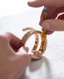 Close-up of hands using a precision screwdriver to assemble a rose gold diamond-embellished serpent bracelet watch on a light surface.