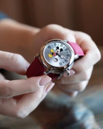 Close-up of hands holding a silver wristwatch with a Mickey Mouse graphic dial and a dark pink fabric strap