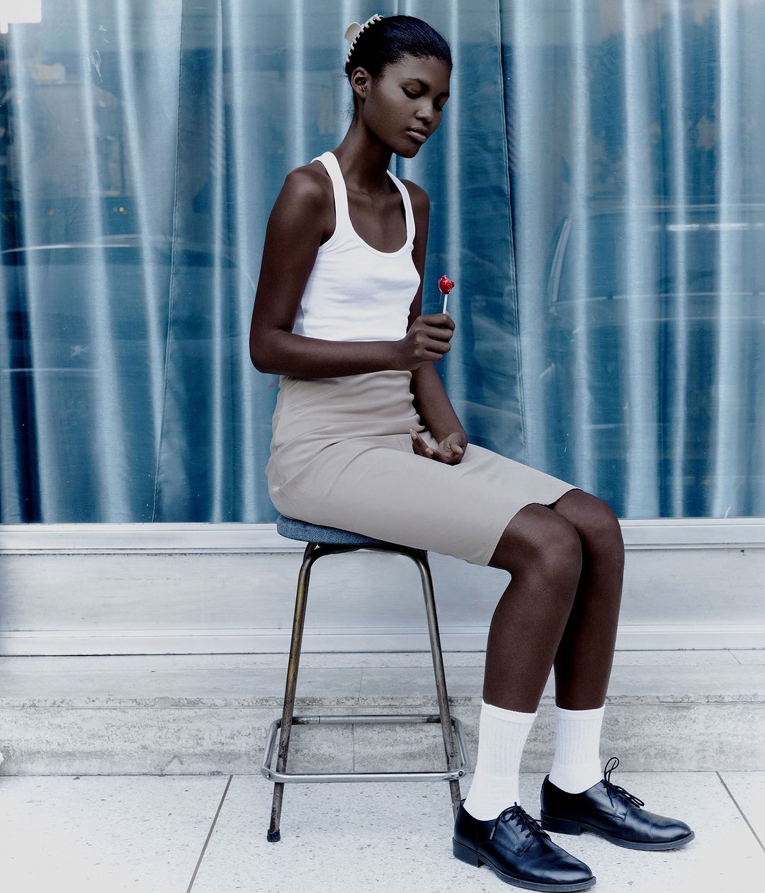 Young woman sits on a stool with lollipop in hand, wearing a white tank top, beige skirt, socks and black dress shoes, against a blue backdrop.