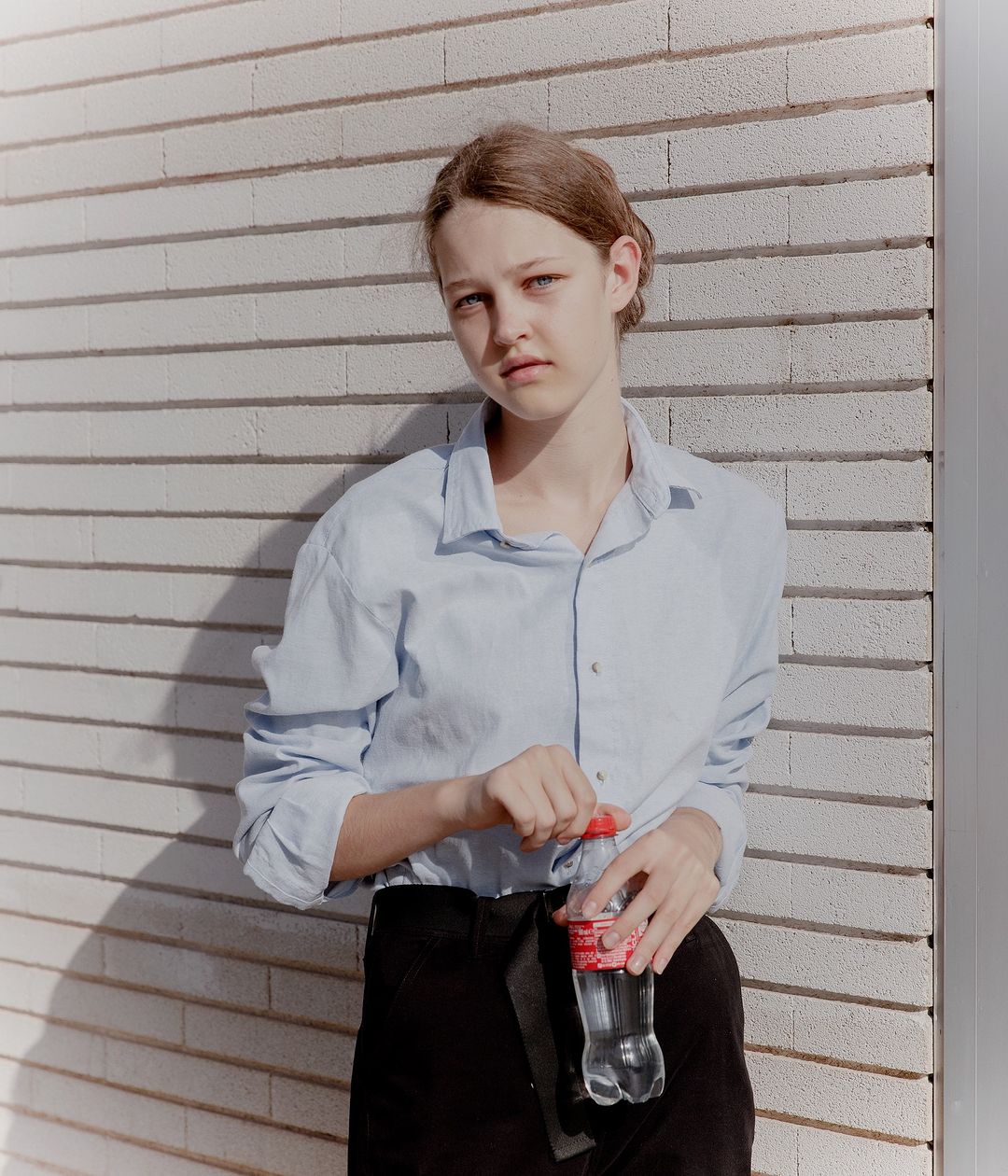 Young woman in casual attire stands by brick wall holding a water bottle. The photo captures natural light and a calm mood.