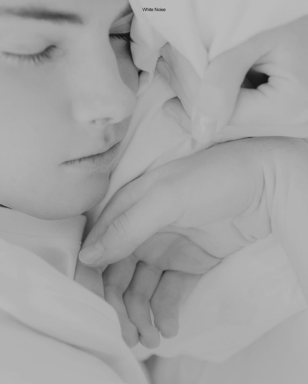 Monochromatic studio portrait of a sleeping young woman with closed eyes, her face partially covered by a white cloth, soft lighting.