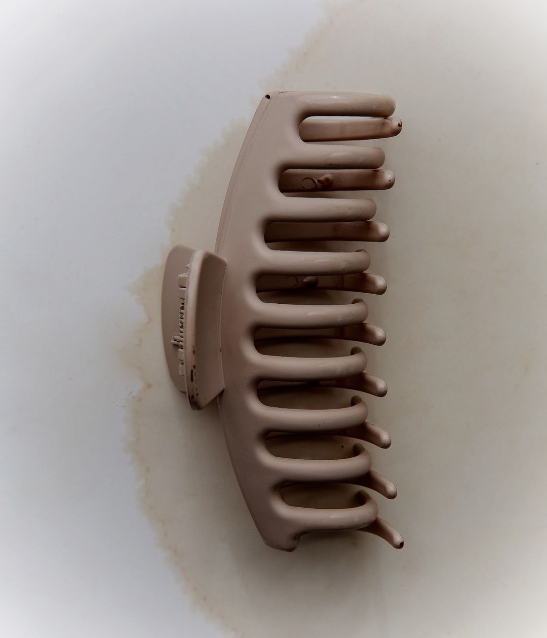 Overhead shot of a beige-colored claw clip with rounded edges and comb-like teeth lying on a white surface with a shadow.