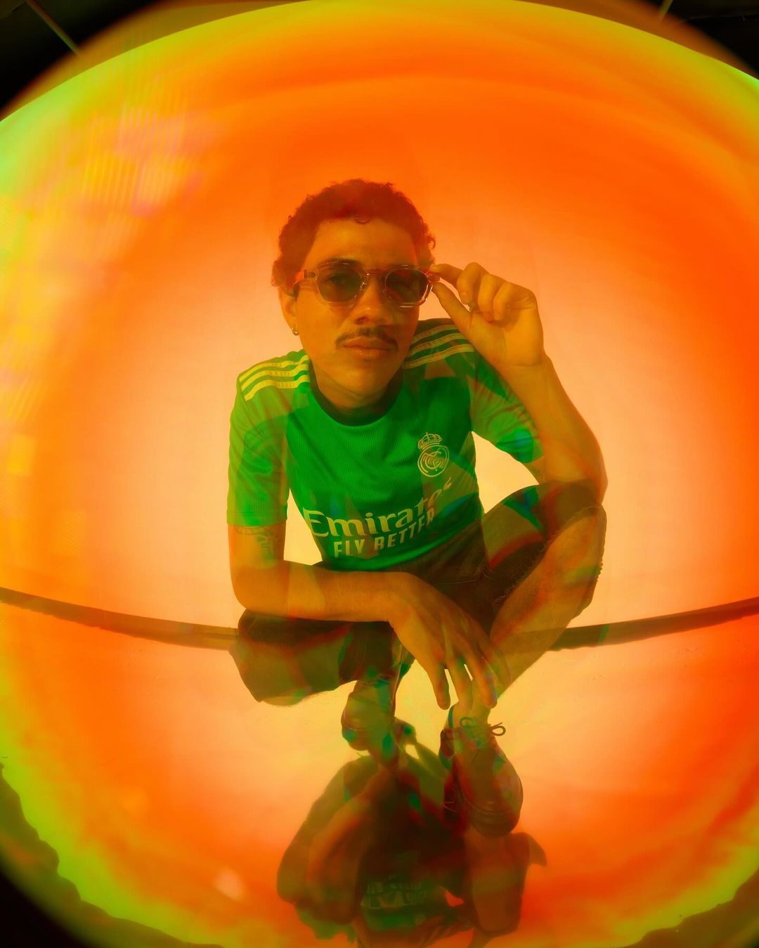 Man with mustache and glasses poses in a green soccer jersey, crouching against a vibrant orange and green backdrop, captured with a fisheye lens.