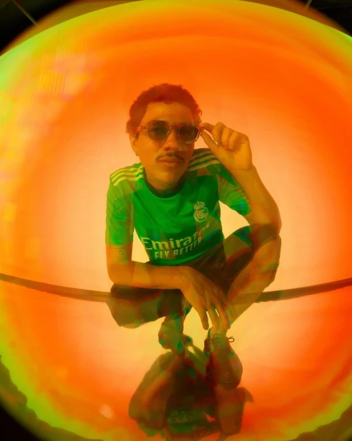 Man with mustache and glasses poses in a green soccer jersey, crouching against a vibrant orange and green backdrop, captured with a fisheye lens.