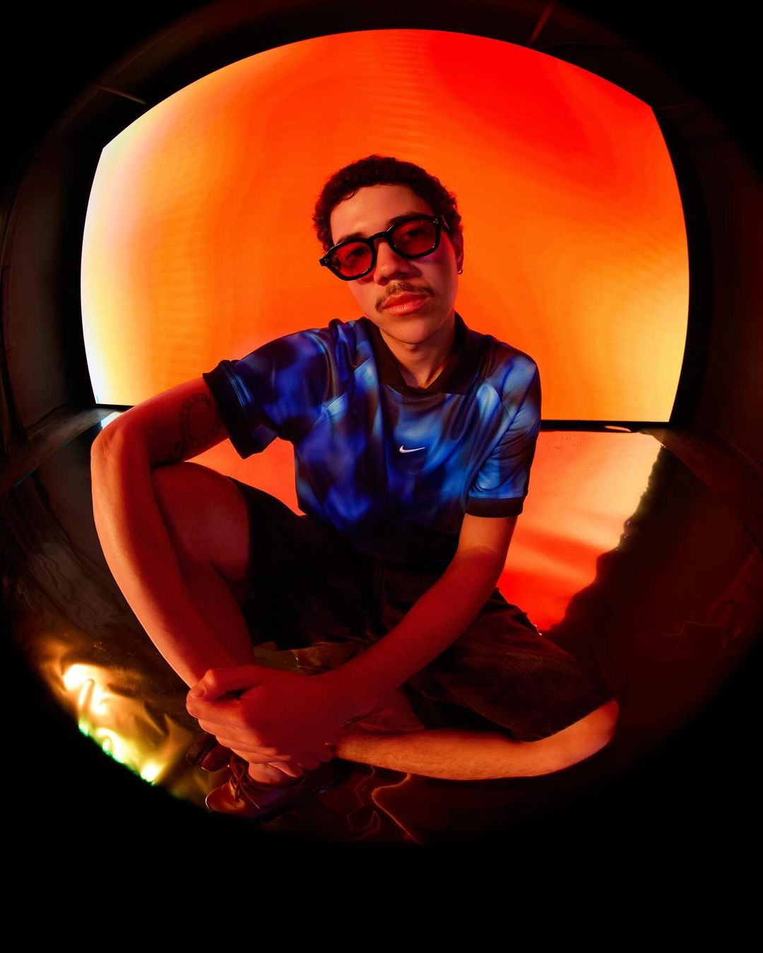 Portrait of a young man in blue shirt and sunglasses, posing in front of a red background with a fisheye lens.