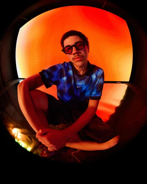 Portrait of a young man in blue shirt and sunglasses, posing in front of a red background with a fisheye lens.