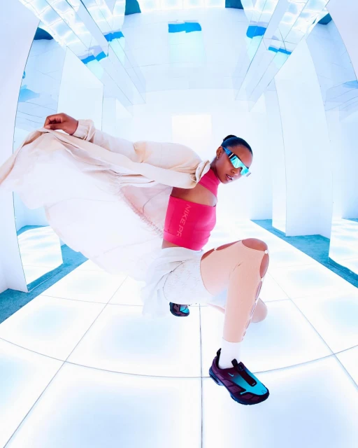 Athletic woman in pink top, light jacket, and futuristic sunglasses poses in a bright, futuristic-looking white hallway while kneeling on the floor.