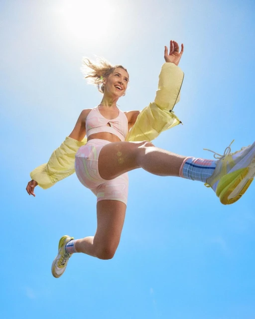 Active woman mid-air, jumping against a vibrant blue sky, wearing workout attire, sneakers and a lightweight jacket, joyful expression.