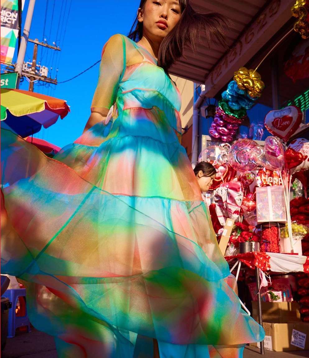 Asian woman in a vibrant watercolor-print dress stands outside a shop in an urban setting, balloons and street signs visible.