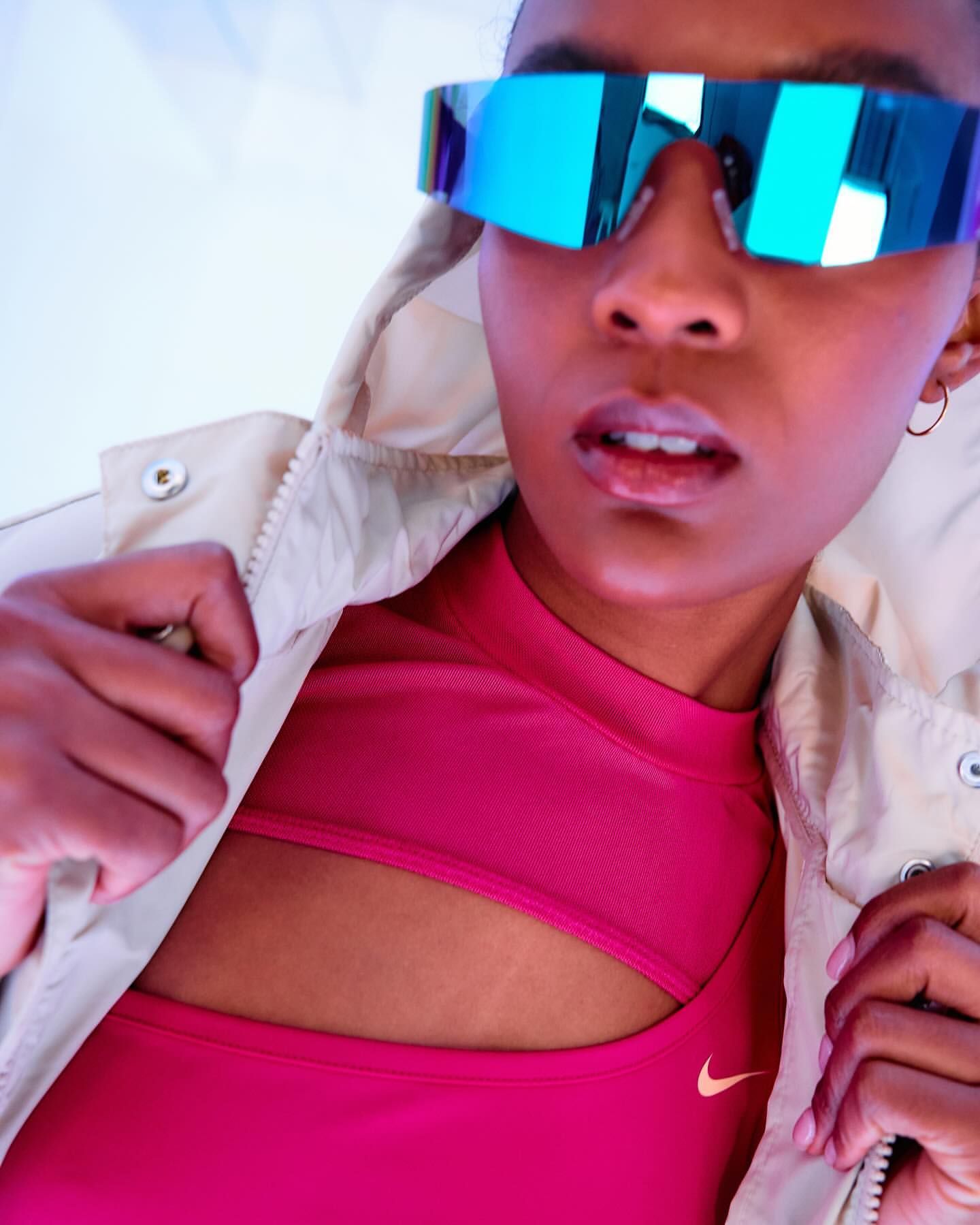 Woman wearing oversized iridescent sunglasses and a fuchsia top and jacket in a brightly lit indoor setting, fashion and portrait photography.