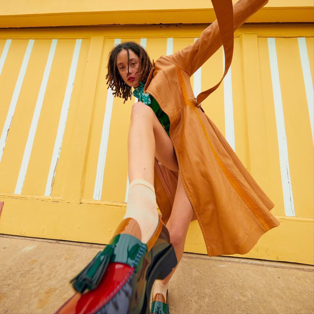 Woman in a brown trench coat and colorful shoes, posing against a yellow wall with white stripes, low angle. Fashion editorial.