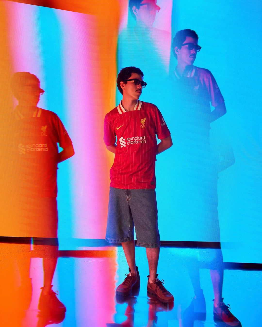 Young man in a red jersey and denim shorts standing against a vibrant multi-colored light projection backdrop by Gabriel Mendoza Weiss.