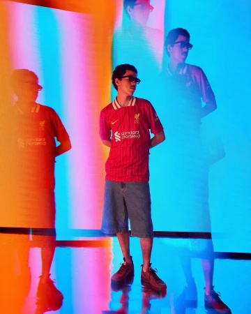 Young man in a red jersey and denim shorts standing against a vibrant multi-colored light projection backdrop by Gabriel Mendoza Weiss.