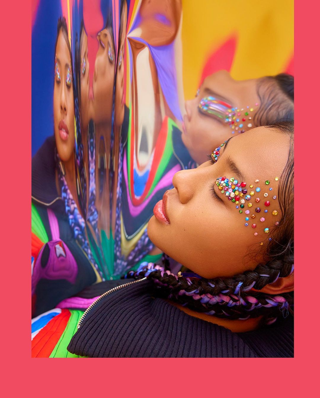 Close-up of a woman with jeweled eye makeup, reflecting in a distorted mirror, wearing a black zip-up jacket, against a colorful background.