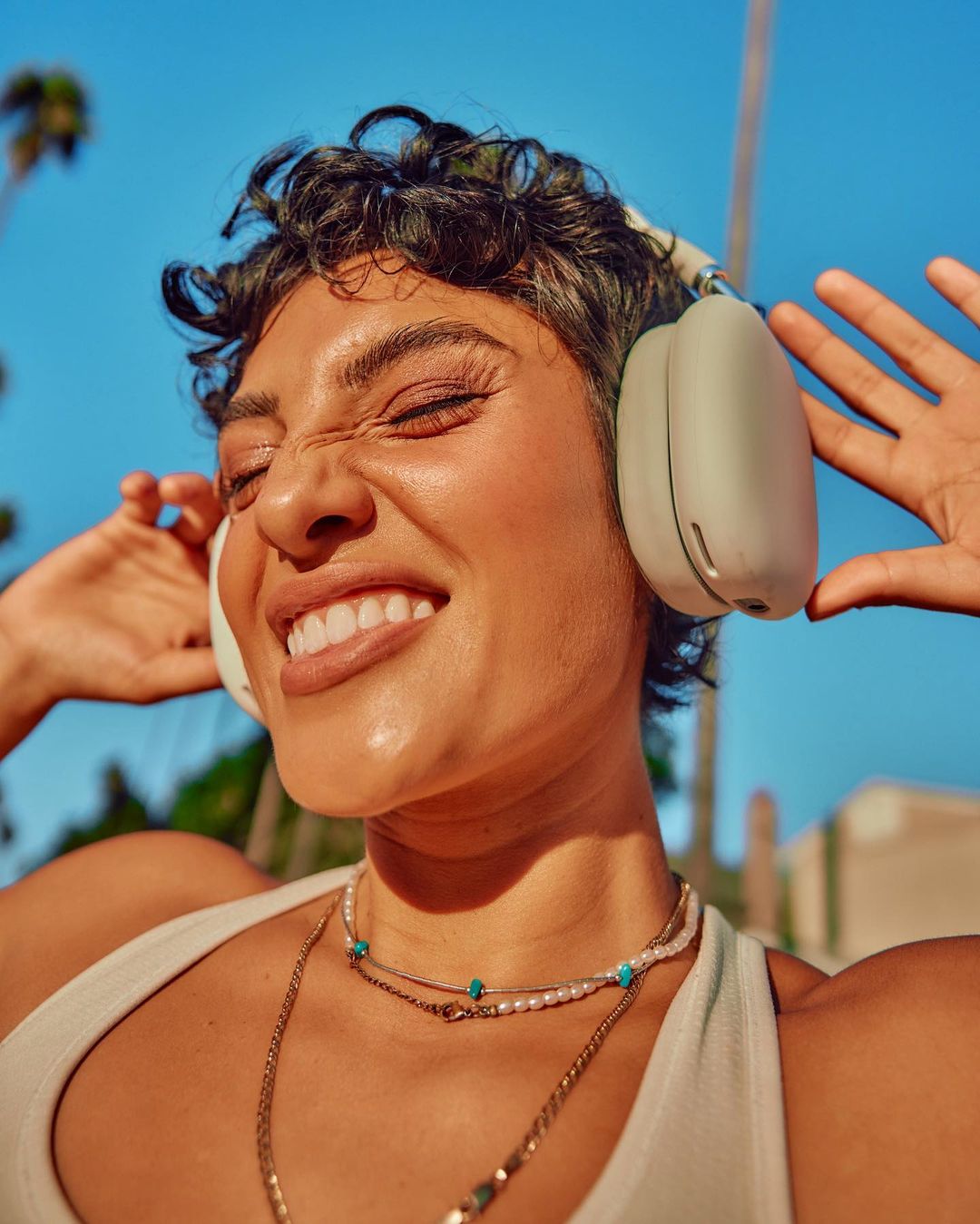 A cheerful woman with headphones is radiantly smiling outdoors in bright sunlight, wearing jewelry and a tank top against a blue sky.