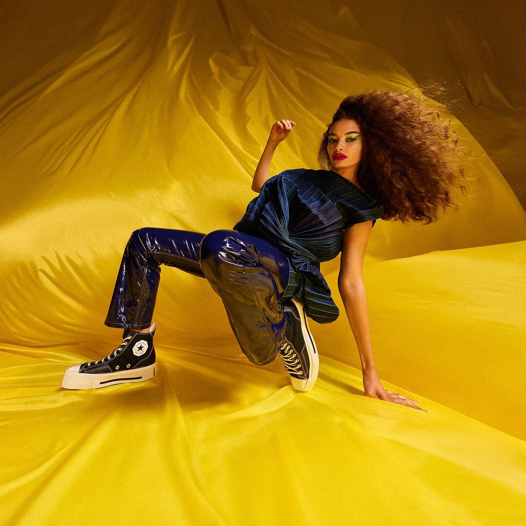 A woman with afro wearing patent leather pants and sneakers poses against a draped yellow textile backdrop in a studio shot.