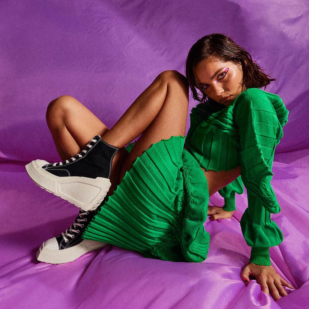 Brunette woman poses in a green, pleated matching set and black high-top sneakers against a light purple backdrop in a studio setting.