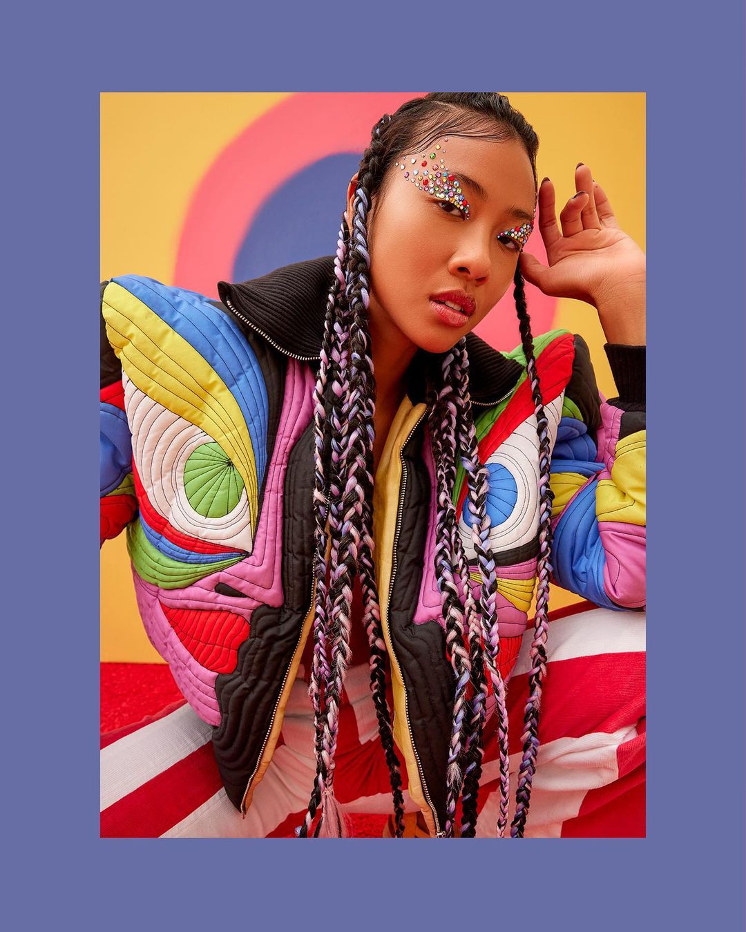 Stylish woman with braided hair and jeweled makeup wears a colorful, geometric-patterned jacket against a vibrant yellow backdrop.