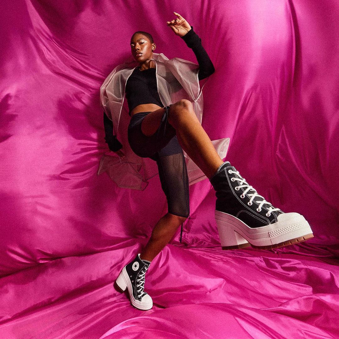 Fashion shot of a model posing on pink fabric background, wearing black shirt, bike shorts, and black platform high-top sneakers.