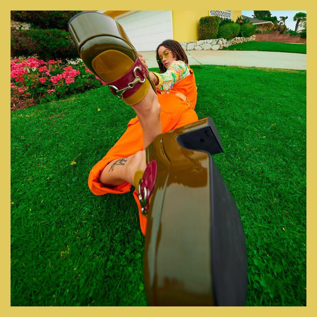 Woman in orange jumpsuit poses on grass lawn with olive green platform shoes, exterior shot. Editorial fashion photograph, low angle.