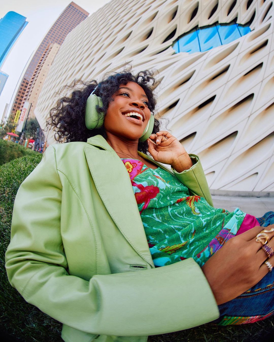 Smiling African-American woman in a green jacket and headphones lies on a grassy lawn with modern architecture in the background.