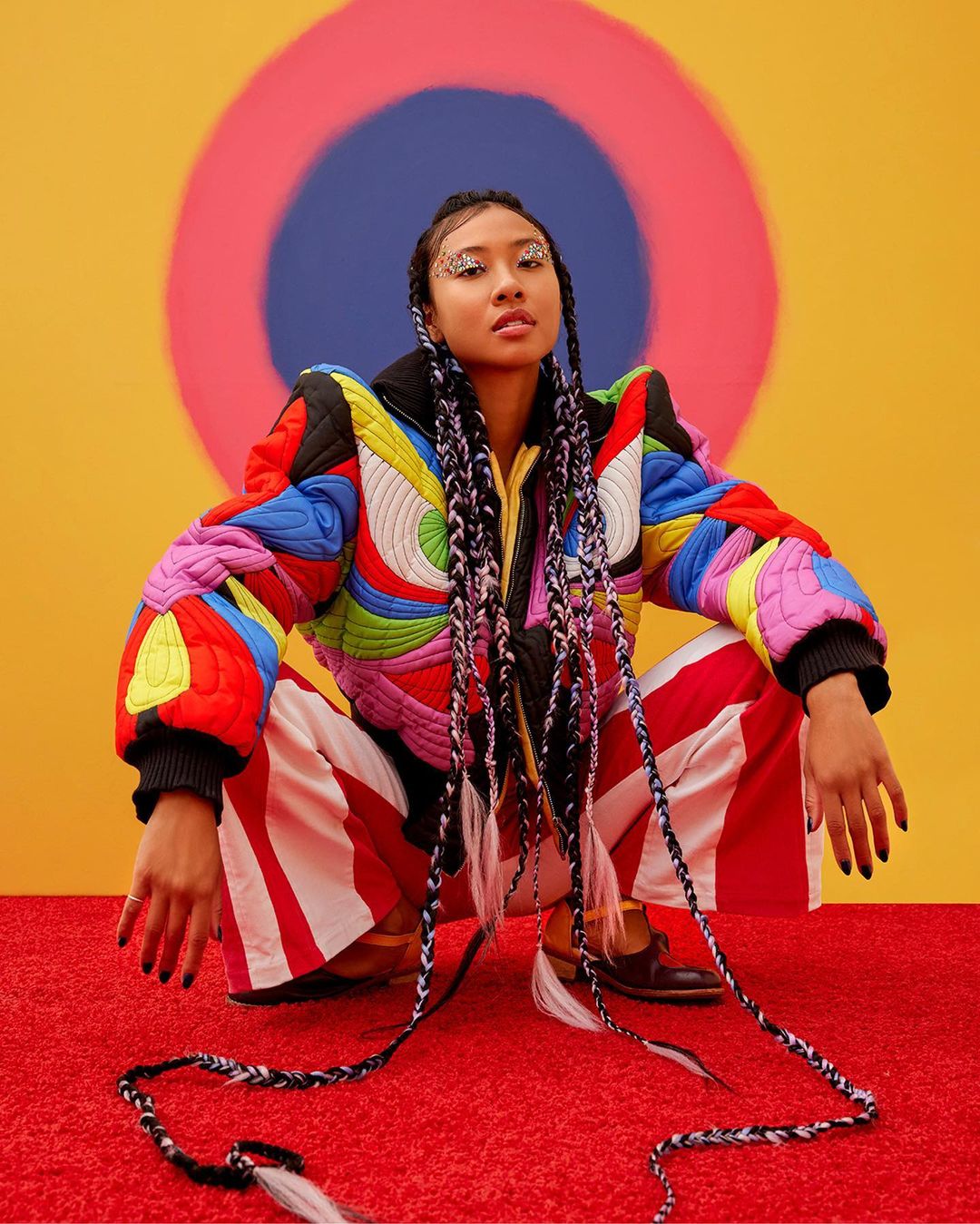 Woman in colorful jacket and red and white striped pants poses against a yellow backdrop with a target graphic, looking at camera.