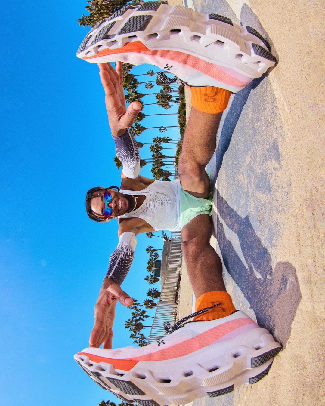 Athletic man poses with running shoes on a beach promenade, blue sky, palm trees, and urban surroundings on a sunny day.