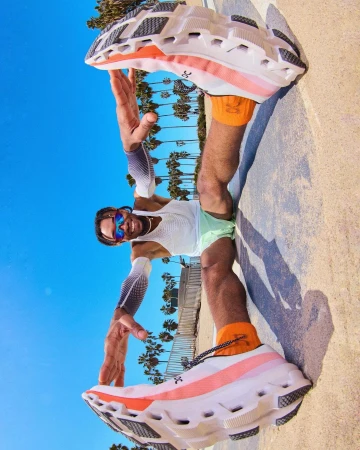 Athletic man poses with running shoes on a beach promenade, blue sky, palm trees, and urban surroundings on a sunny day.