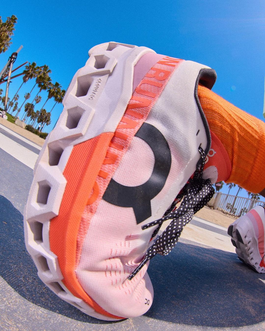 Close-up of an 'On' brand running shoe with a white and coral sole, black logo, and orange sock, taken outdoors on a sunny day.