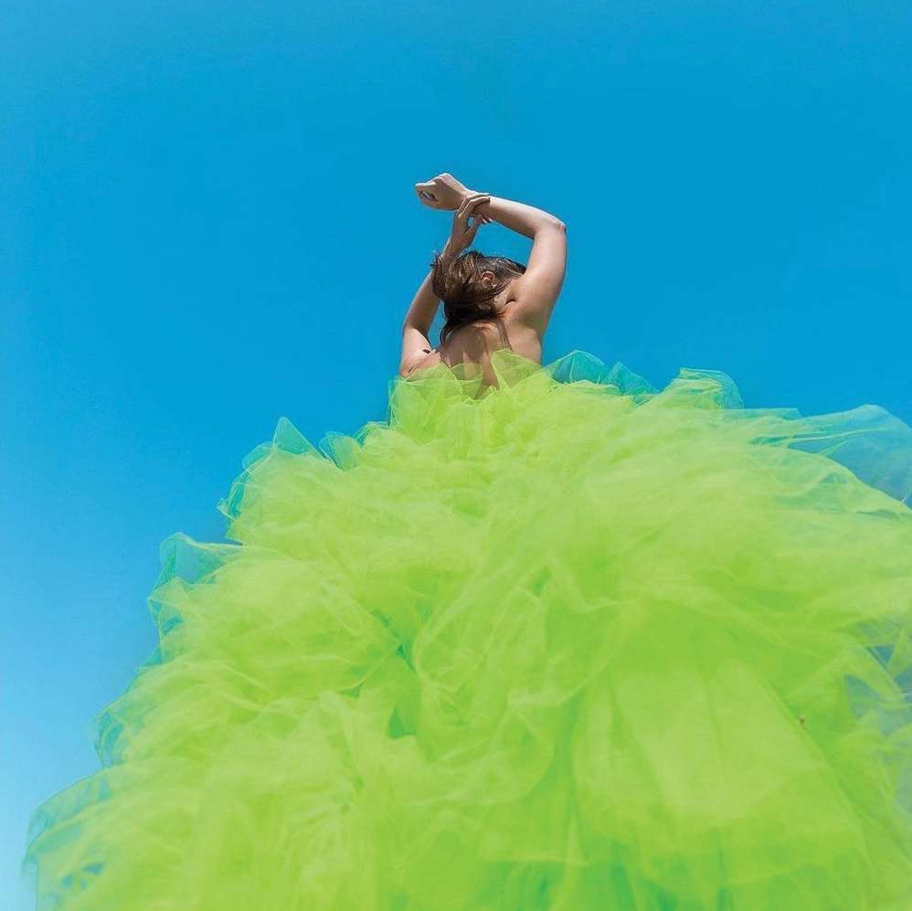 A woman in a voluminous chartreuse gown, viewed from the back, stands against a clear blue sky with her arms slightly raised.