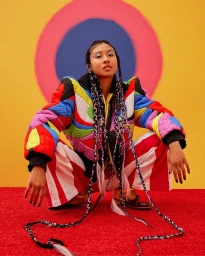 A woman crouches against a bright yellow wall with concentric pink and blue circles, wearing a vibrant multicolored puffer jacket and striped pants.