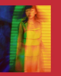 Motion blurred portrait of a woman in a layered dress against a ribbed wall with blue, green, and yellow colored light projections.