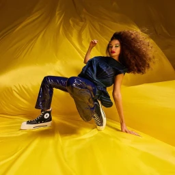 Model with voluminous curly hair wearing dark patent blue trousers and platform sneakers on a rippled yellow backdrop