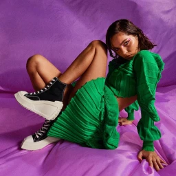 Woman in a green pleated skirt and matching top wearing black platform high-top sneakers against a purple draped backdrop.
