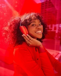 Black woman wearing red headphones and a red top smiling while holding her headphones against a vibrant red backdrop.