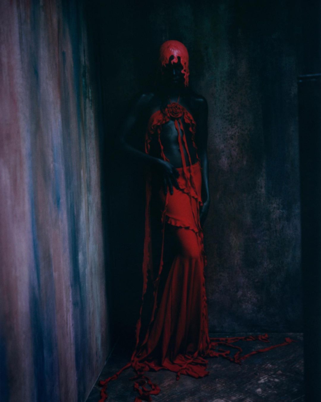 Person in red dress and matching head covering stands in a dimly lit room with textured walls, creating a surreal and dramatic visual.